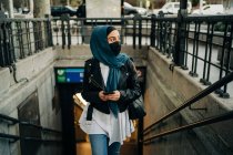 Этническая женщина в хиджабе и защитной маске, идущая наверх по городской улице, используя смартфон и отворачиваясь — стоковое фото