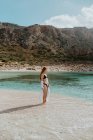 Piena lunghezza sottile femminile in costume da bagno nero in piedi sulla spiaggia di sabbia Balos contro scogliera rocciosa e guardando lontano sul chiaro tempo soleggiato — Foto stock