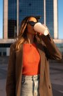 Молодая женщина в стильной одежде, пьет горячий напиток к солнцезащитным очкам в городе — стоковое фото