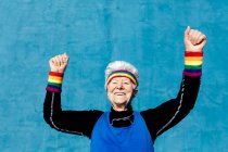 Excited senior female in sportswear celebrating victory with clenched fists and raised arms on blue background in studio — Stock Photo