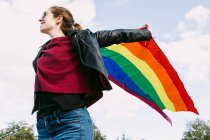 Desde abajo encantada lesbiana de pie en la calle con la bandera del arco iris LGBT ondeando en el viento y mirando hacia otro lado contra el cielo nublado - foto de stock