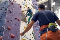 From below of brave female athlete climbing artificial wall in bouldering center under supervision of professional instructor — Stock Photo