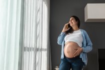 Smiling pregnant female touching belly while standing in room at home and talking mobile phone — Stock Photo