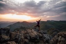 Vue arrière de l'homme bondissant sur les rochers du haut sommet de la crête de montagne sous un ciel nuageux au coucher du soleil — Photo de stock