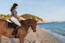 Side view of young female looking away while riding chestnut horse on sandy sea shore against mount under light sky — Stock Photo
