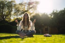 Conteúdo de corpo inteiro jovem do sexo feminino com os olhos fechados vestindo casual desgaste sentado em Lotus Pose em verdejante clareira gramada e meditando no parque ensolarado — Fotografia de Stock