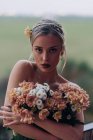 Charming romantic young bare shouldered female with bunch of fresh blooming flowers standing against green field background in a balcony — Fotografia de Stock