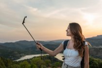 Seitenansicht einer reisenden Frau mit Rucksack, die auf einem Hügel steht und im Sommer Selbstaufnahmen auf dem Smartphone vor dem Hintergrund der Gebirgskette macht — Stockfoto