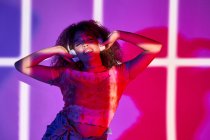 Mujer afroamericana feliz de cuerpo completo en pantalones cortos escuchando música favorita en auriculares y bailando con los ojos cerrados en luces de neón en el estudio - foto de stock