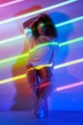 Corpo inteiro elegante confiante dançarina afro-americana com cabelo encaracolado e óculos de sol apoiados na parede olhando para a câmera em luzes de néon no estúdio de dança — Fotografia de Stock