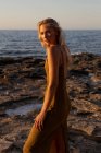 Vista lateral da jovem mulher em pé na costa contra o mar azul acenando ao pôr do sol e olhando para a câmera — Fotografia de Stock