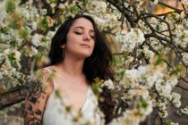 Young female with tattooed arm wearing white dress and standing in flowers of tree with closed eyes — Fotografia de Stock