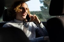 Веселий молодий стильний чоловік сидить на водійському сидінні і розмовляє на мобільному телефоні під час подорожі по природі в сонячний літній день — стокове фото