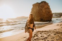 Side view slender young female in black swimwear with long hair standing on sandy seacoast near rough rocks on sunny hot day in Fyriplaka Milos — Stock Photo