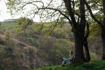 Side view of female traveler with paper guide sitting on meadow between green trees and mount in countryside - foto de stock