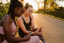 Веселые многорасовые спортсменки в активной одежде, сидящие на скамейке в парке и пользующиеся мобильными телефонами вместе после тренировки на закате — стоковое фото