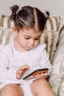 Criança adorável sentada no sofá em casa e assistindo desenhos animados interessantes no telefone móvel — Fotografia de Stock