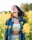 Zufriedene junge Frau in Jeansjacke steht mit geschlossenen Augen auf blühendem, duftendem Rapsfeld an klarem, sonnigem Tag — Stockfoto