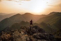 Вид ззаду людини, що стрибає через валуни високого піку гірського хребта під хмарним небом під час заходу сонця — стокове фото