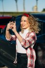 Femme tendance debout près de l'automobile moderne et de prendre selfie sur téléphone mobile — Photo de stock