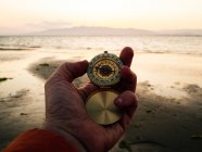 POV crop anonymous adventurer checking route with retro compass while standing on sandy beach near sea at sunset — Fotografia de Stock