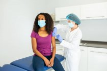 Специалистка-женщина в защитной форме, латексных перчатках и маске для лица, прививающая афроамериканку в клинике во время вспышки коронавируса — стоковое фото