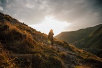 Вид збоку на жінку, що йде вниз по скелястому схилу гори в зеленій долині з хмарним небом заходу сонця на фоні — стокове фото