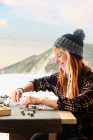 Side view of happy content female traveler creating handmade accessories while sitting at wooden table in parked truck at seaside — Stock Photo
