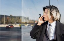 Joven empresario masculino étnico bien vestido con el pelo teñido hablando en el teléfono celular mientras mira hacia otro lado contra la pared de vidrio y el camino urbano - foto de stock