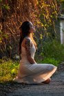 Dreaming lady with tattooed arm wearing white dress and sitting on green lawn in nature — Photo de stock