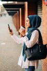 Vista lateral de la mujer étnica en hijab de pie en la plataforma en la estación de tren y tomar selfie en el teléfono móvil mientras espera el tren - foto de stock