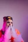 Coole Frau im Streetstyle-Outfit, die E-Zigarette raucht und Rauch durch die Nase auf lila Hintergrund im Studio mit rosa Neonbeleuchtung ausatmet — Stockfoto