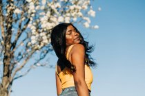 Baixo ângulo de mulher afro-americana sonhadora em pé no parque de primavera florescente e desfrutando de tempo ensolarado com olhos fechados — Fotografia de Stock