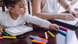 Crop unrecognizable woman browsing laptop while little child sitting at table and drawing with markers in notebook — Stock Photo