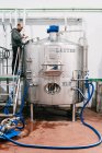 Вид сбоку на мужчину-инженера, осматривающего бак из нержавеющей стали со шлангами в пивоварне с мокрым полом — стоковое фото