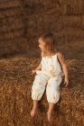 Беззаботный ребенок в комбинезоне сидит на соломенном тюке в солнечный день в сельской местности — стоковое фото