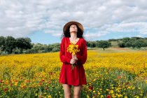 Female in hat with eyes closed holding blossoming yellow flowers in countryside field under cloudy sky — Stock Photo