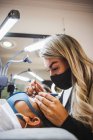 Cosmetologist with tweezers applying fake eyelashes for extension on eye of ethnic client with face protective mask in salon during coronavirus pandemic — Foto stock