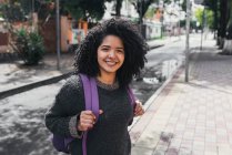 Side view of delighted ethnic female student with Afro hairstyle and backpack standing on street on sunny day and looking at camera — Fotografia de Stock