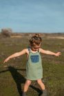 Очаровательная маленькая девочка в комбинезоне, стоящая с протянутыми руками на лугу и смотрящая вниз — стоковое фото