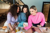 Sonrientes amigas jóvenes que usan ropa casual navegando por teléfonos móviles mientras se reúnen para almorzar en el restaurante - foto de stock