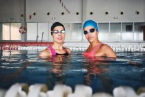 Femmes sportives en bonnets et maillots de bain se préparant pour l'entraînement dans la piscine avec de l'eau transparente pendant la journée — Photo de stock