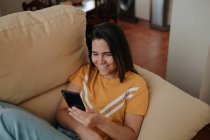 Mensajería de texto femenina joven en el teléfono celular mientras está acostado en el sofá en la sala de estar - foto de stock