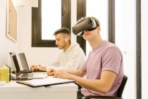 Side view of positive young male employee in casual clothes experiencing virtual reality in modern headset while working on laptop with smiling ethnic colleague — Fotografia de Stock