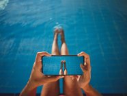 From above of crop anonymous barefoot female taking photo of legs on cellphone over swimming pool with pure water — Stock Photo