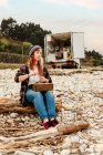 Анонимная ремесленница, создающая бижутерию ручной работы, используя полудрагоценные камни и сидя на пляже у моря — стоковое фото