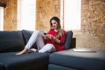 Female remote employee text messaging on cellphone while sitting on couch against tablet in loft style house — Photo de stock
