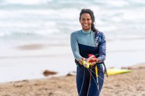 Happy ethnic female kiter in wetsuit with kitesurfing equipment looking at camera on sandy ocean beach — Stock Photo