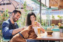 Casal étnico alegre com copos de cerveja e batatas fritas falando contra o cão de raça pura à mesa à luz do sol — Fotografia de Stock