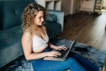 Side view of young self employed woman in casual clothes and eyeglasses sitting on the floor near comfortable sofa and working remotely on laptop — Stock Photo